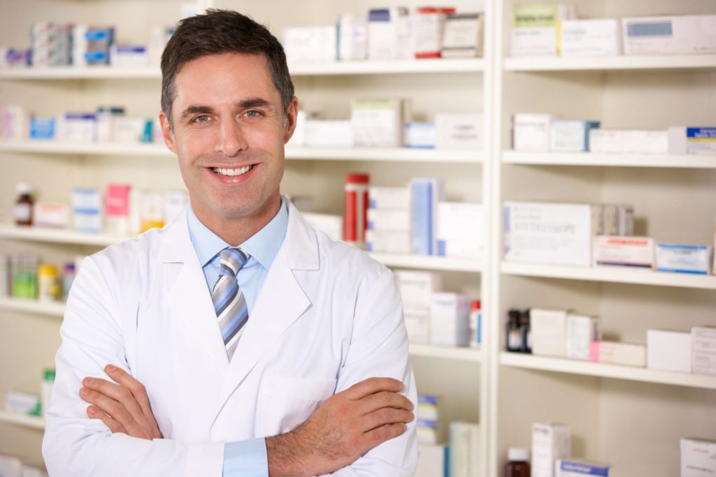 A man in white lab coat standing next to shelves of medicine.